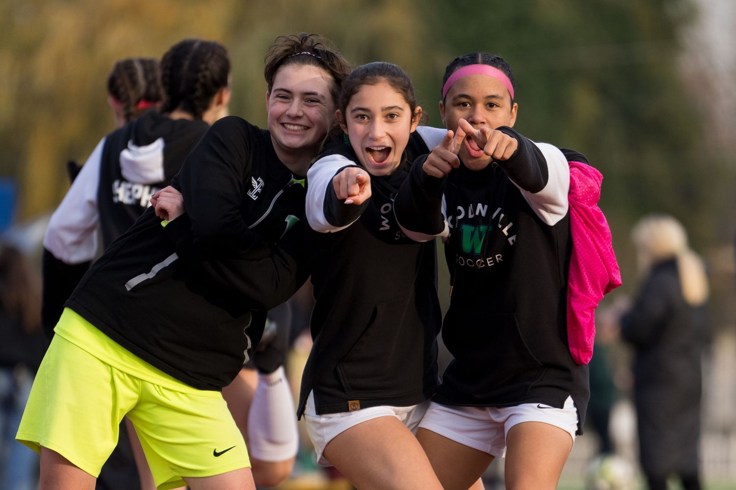 A group of athletes in athletic wear point and smile at the camera during an outdoor sports event.