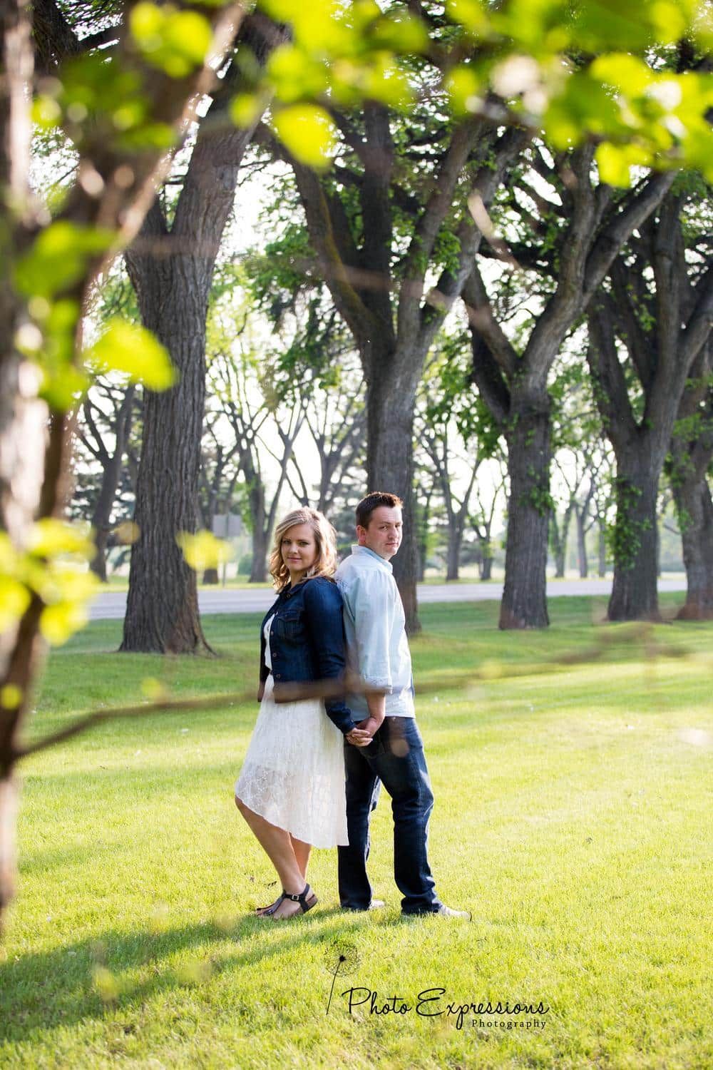 Couple walking through park with tall trees and lush grass in summer.