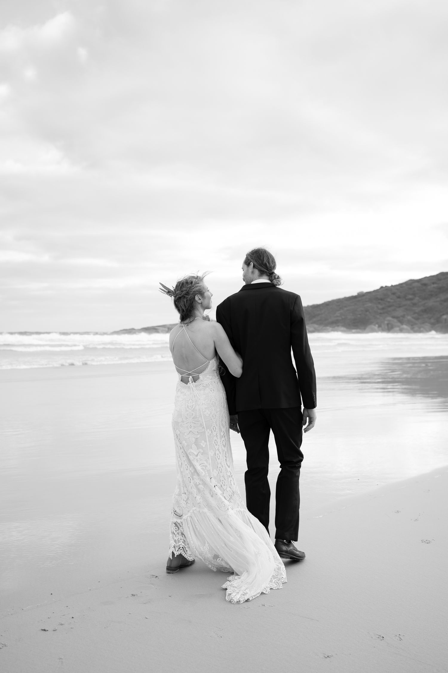 Black and white photo of newlyweds walking along a beach, bride in lace dress and groom in dark suit.