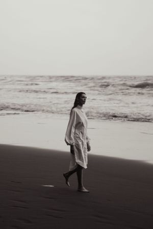 Monochrome image of person walking along misty beach in flowing white dress.