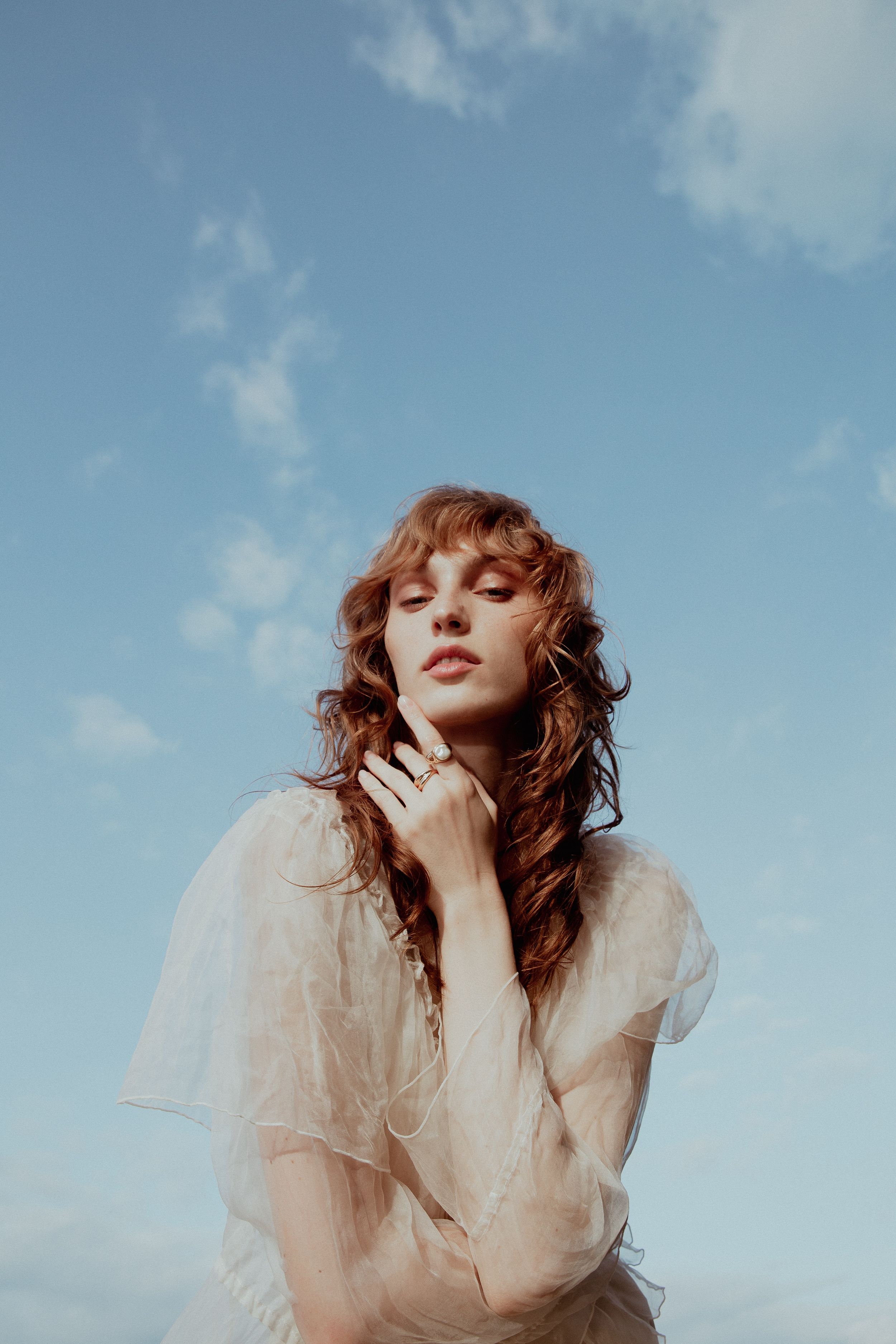 Ethereal portrait of woman in sheer white top against blue sky background.