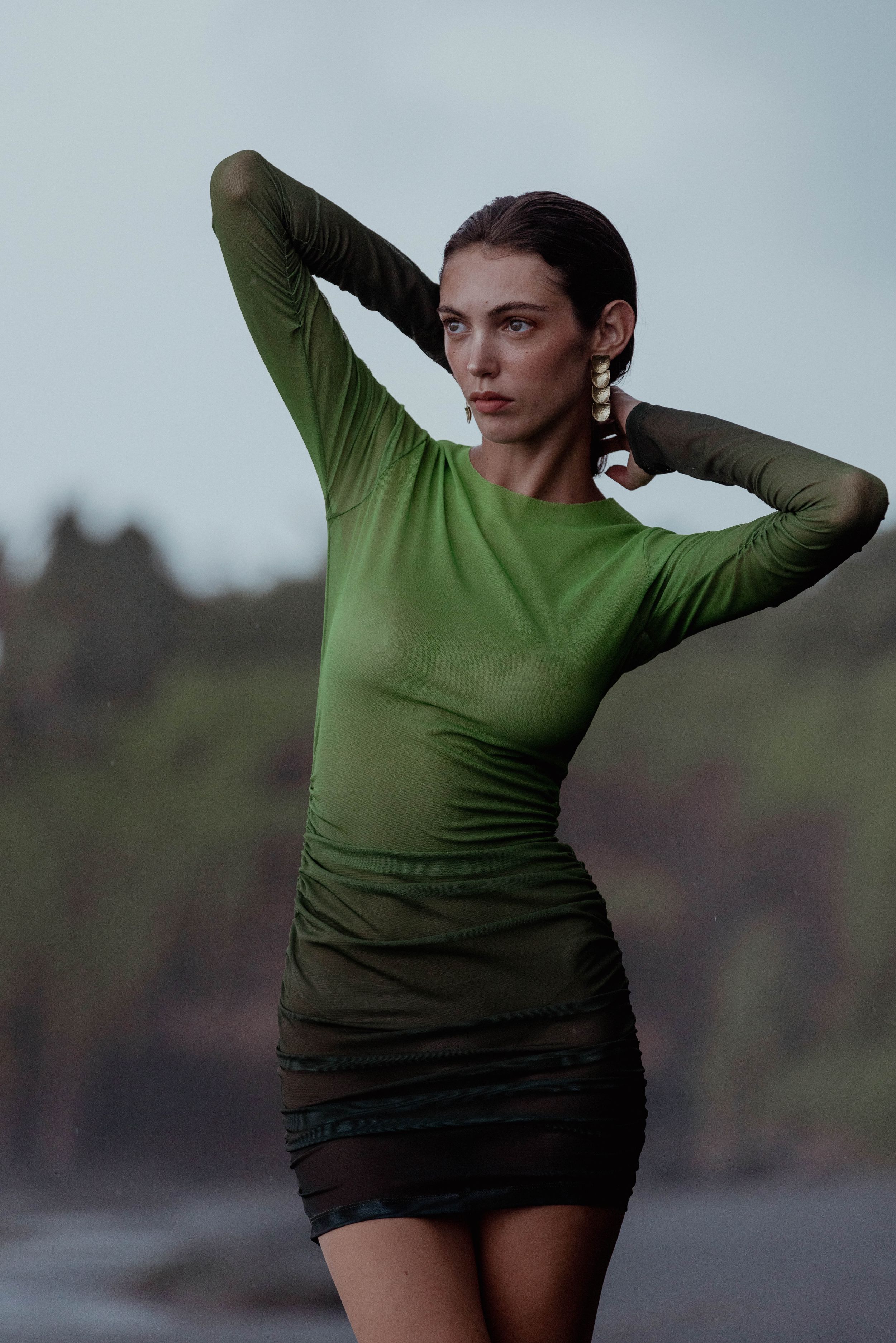 Fashion model in form-fitting green ombre dress poses against moody coastal backdrop.