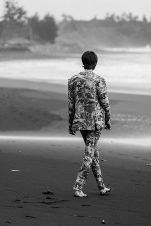 Black and white beach scene with figure in patterned suit walking on sand.