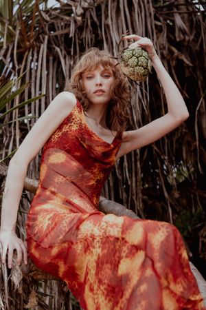 Fashion shot of model in red flowing dress holding pineapple against tropical backdrop.