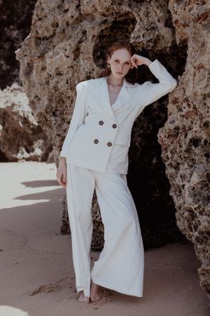 Woman in crisp white suit posing in front of rocky cave entrance in fashion editorial.