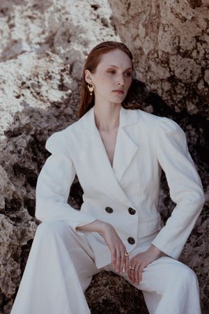 Model in elegant white pantsuit sitting against rocky backdrop in professional photoshoot.