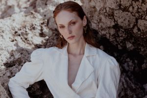 Fashion model wearing white blazer posed against textured stone wall in artistic portrait.