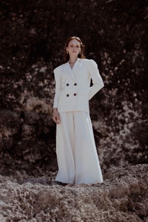 Full-length fashion photo of model in elegant white pantsuit on rocky terrain.