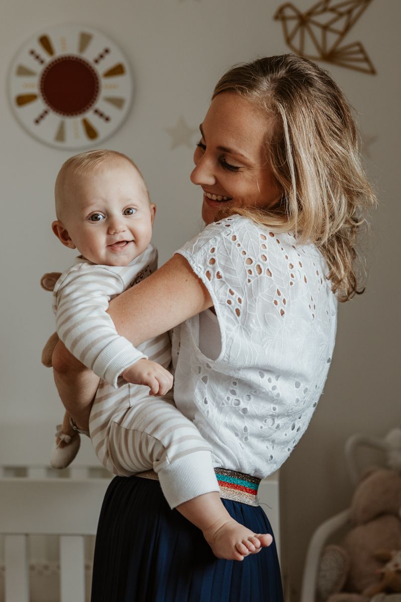 maman joyeuse portant son bebe souriant dans les bras pendant une seance photo de famille a domicile a Lille