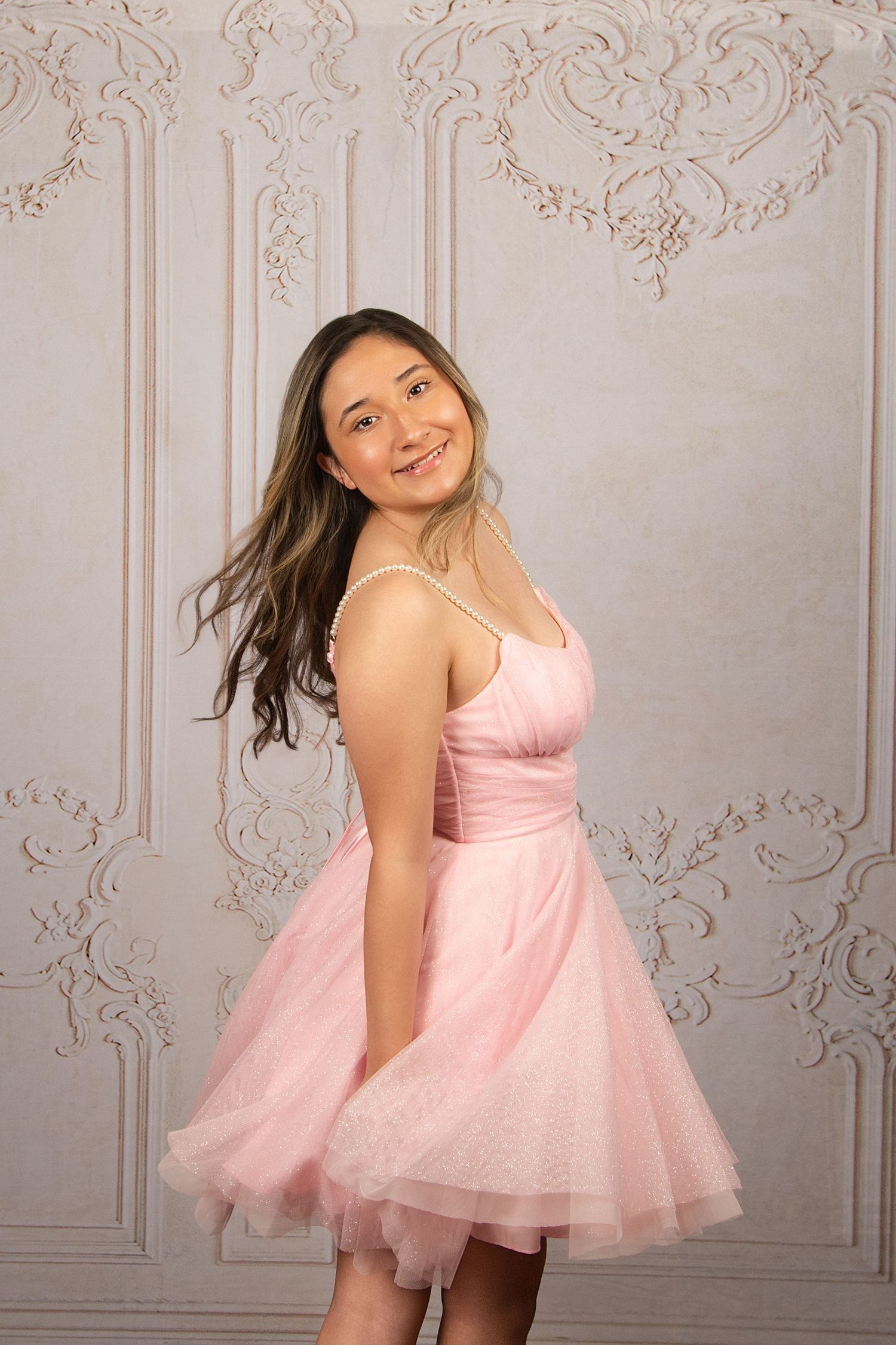 Young woman smiling in a pink tulle dress with pearl necklace, posing against an ornate white wall.