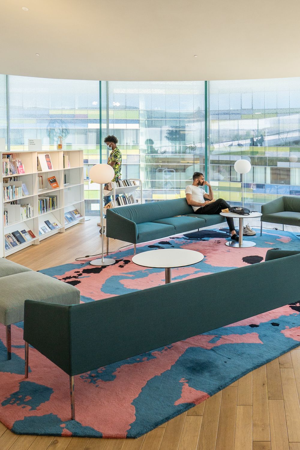 Modern library lounge with green sofas, geometric rug, and floor-to-ceiling windows overlooking cityscape.