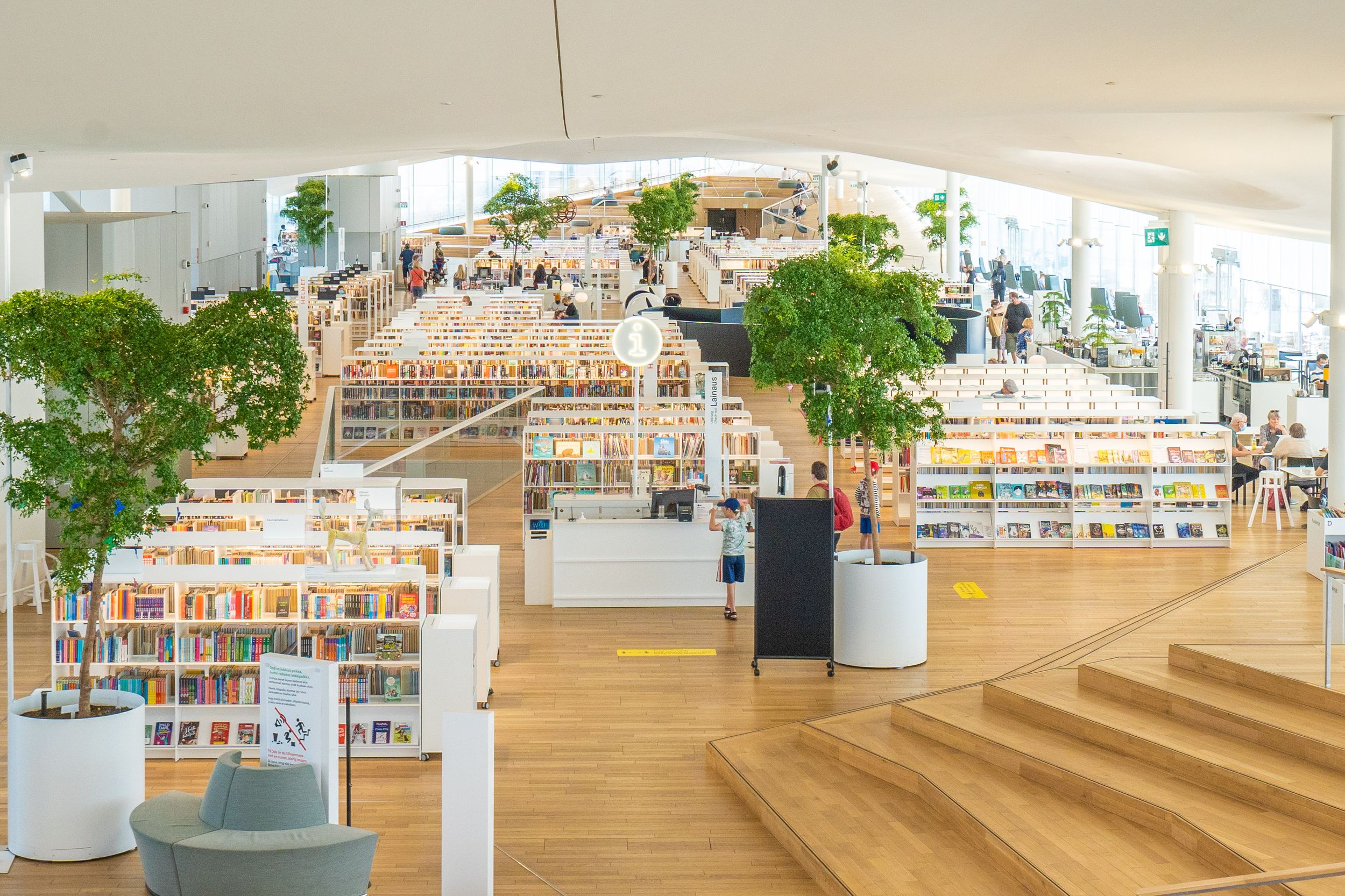 Modern library interior with wooden tiered seating and bookshelves arranged in an open-plan design under natural lighting.