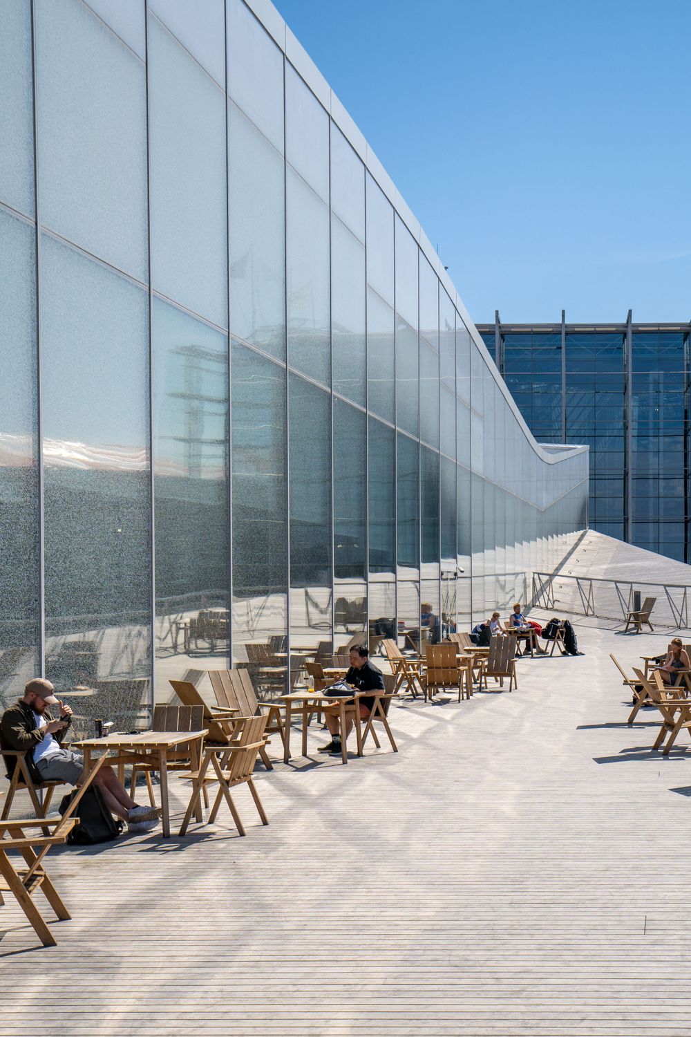 Outdoor terrace with wooden chairs along a curved glass wall on a sunny day.