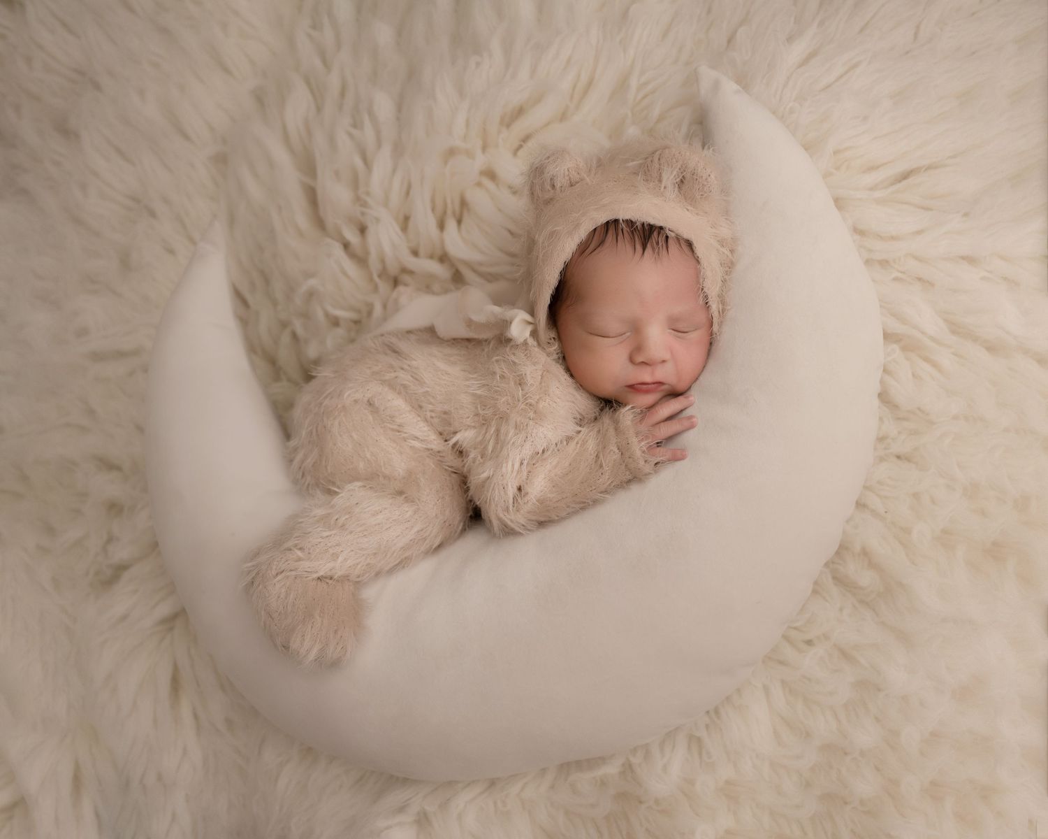 A newborn dressed in a fuzzy beige outfit sleeps peacefully in a white crescent-shaped prop on soft fur.