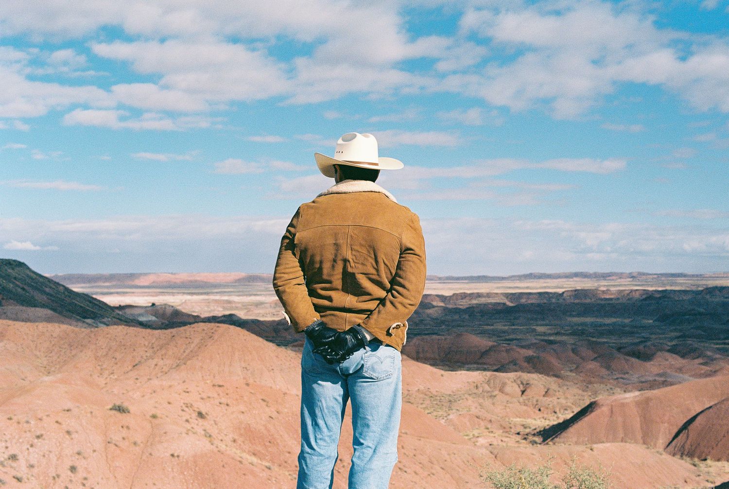Person in brown jacket and jeans overlooking a vast desert landscape under blue skies.