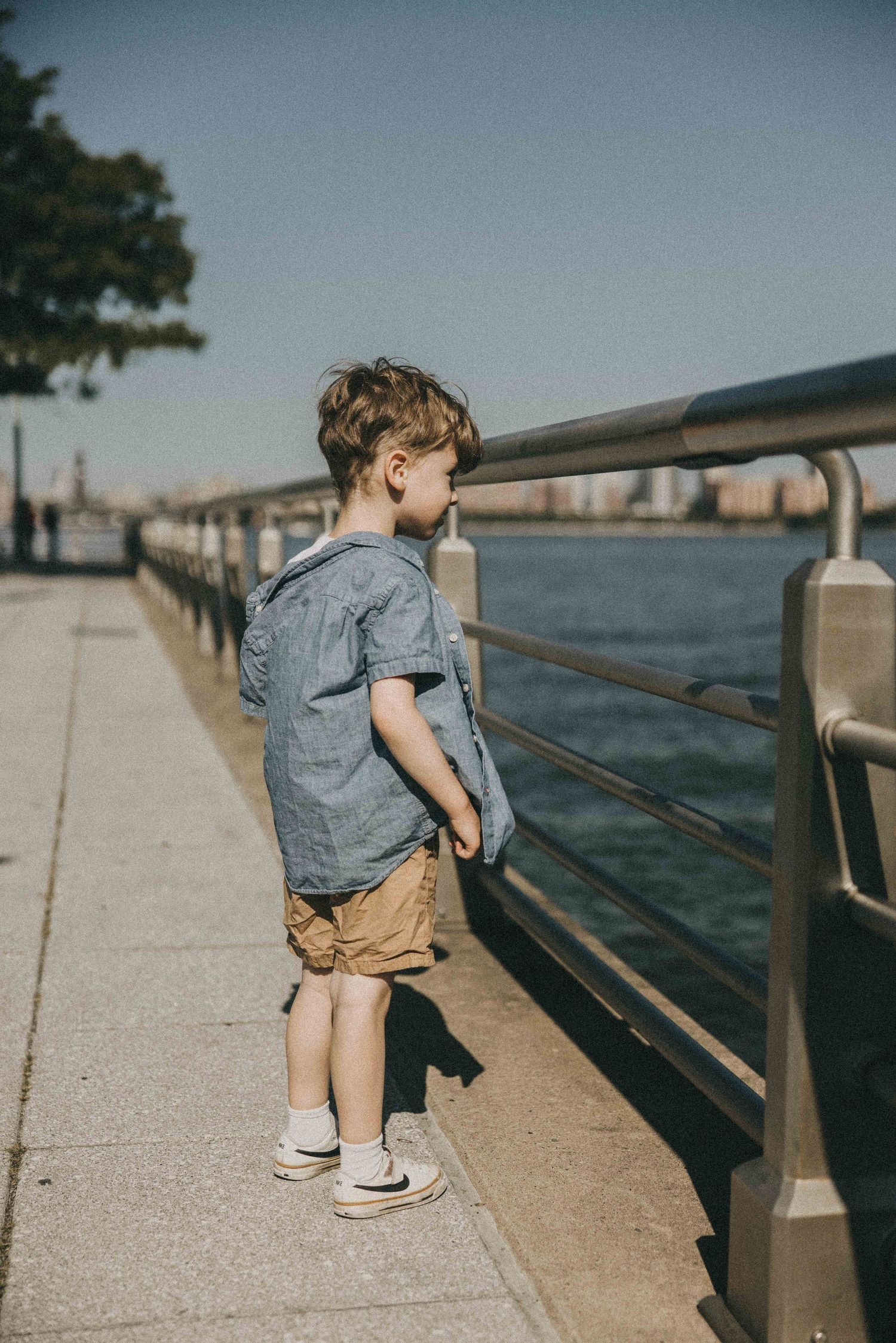 Small child exploring waterfront boardwalk wearing denim and brown backpack on sunny day.