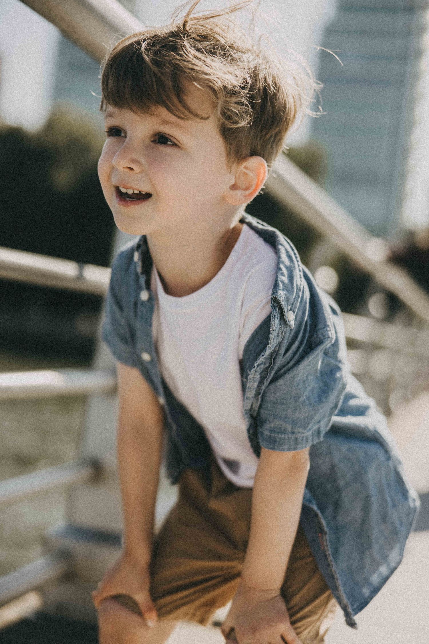 Small child wearing a denim shirt and brown pants laughs joyfully while playing outdoors on a sunny day.