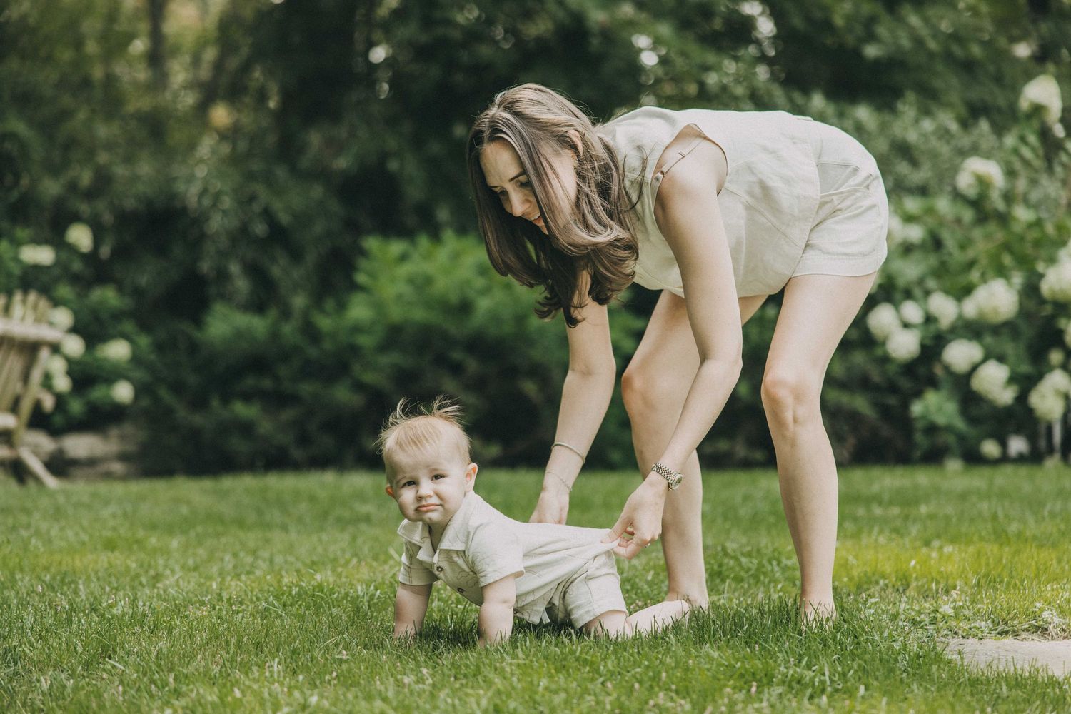 An adult and baby play together on a sunny lawn surrounded by flowering bushes and greenery.