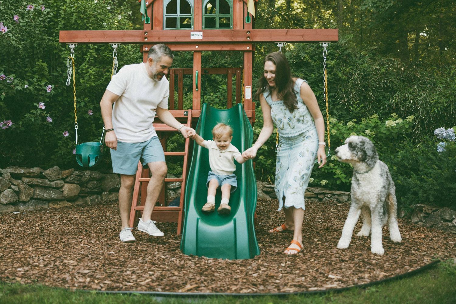 Family with baby on green slide at backyard wooden playset, large gray dog standing nearby on mulch.