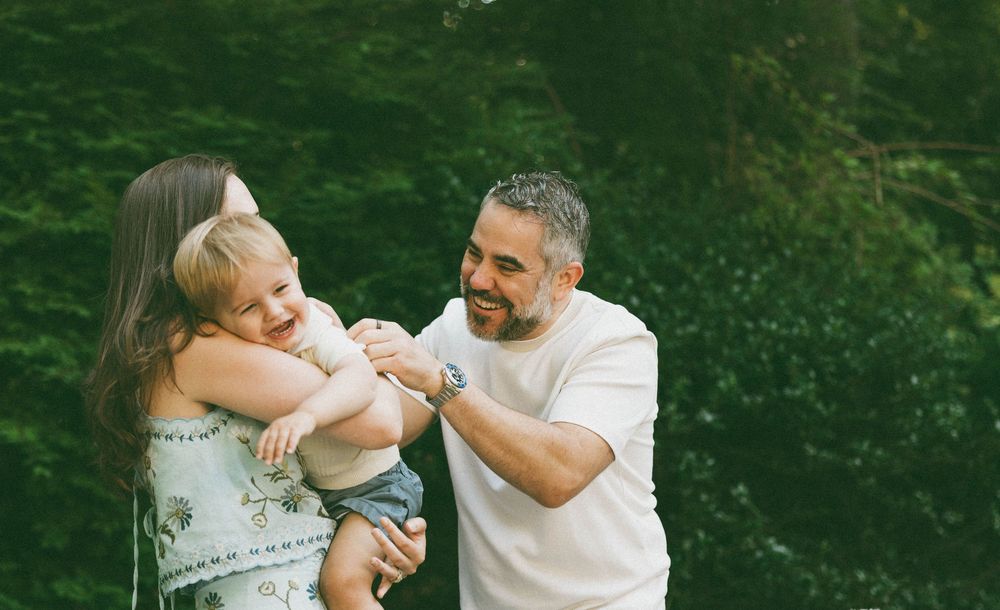 A happy family with a mother, father, and toddler smiling outdoors surrounded by lush green trees.