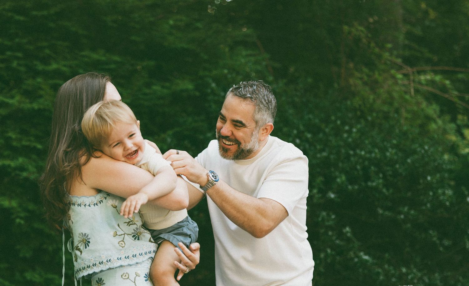 A happy family with a mother, father, and toddler smiling outdoors surrounded by lush green trees.