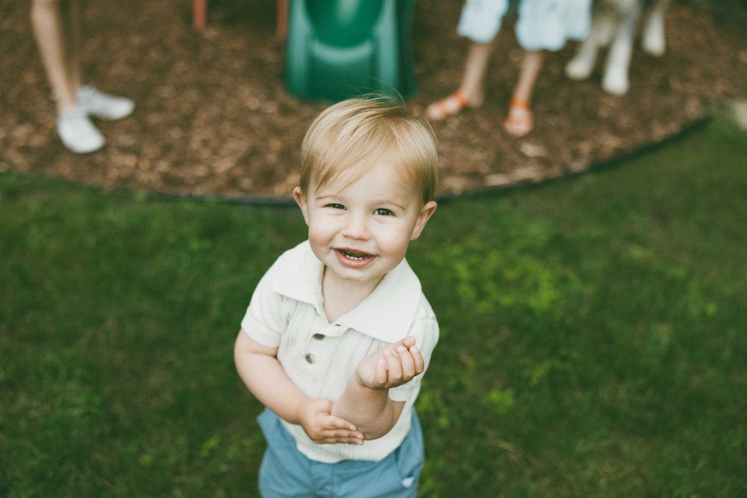 Smiling toddler boy with blonde hair standing on grass near a playground, looking up at the camera.