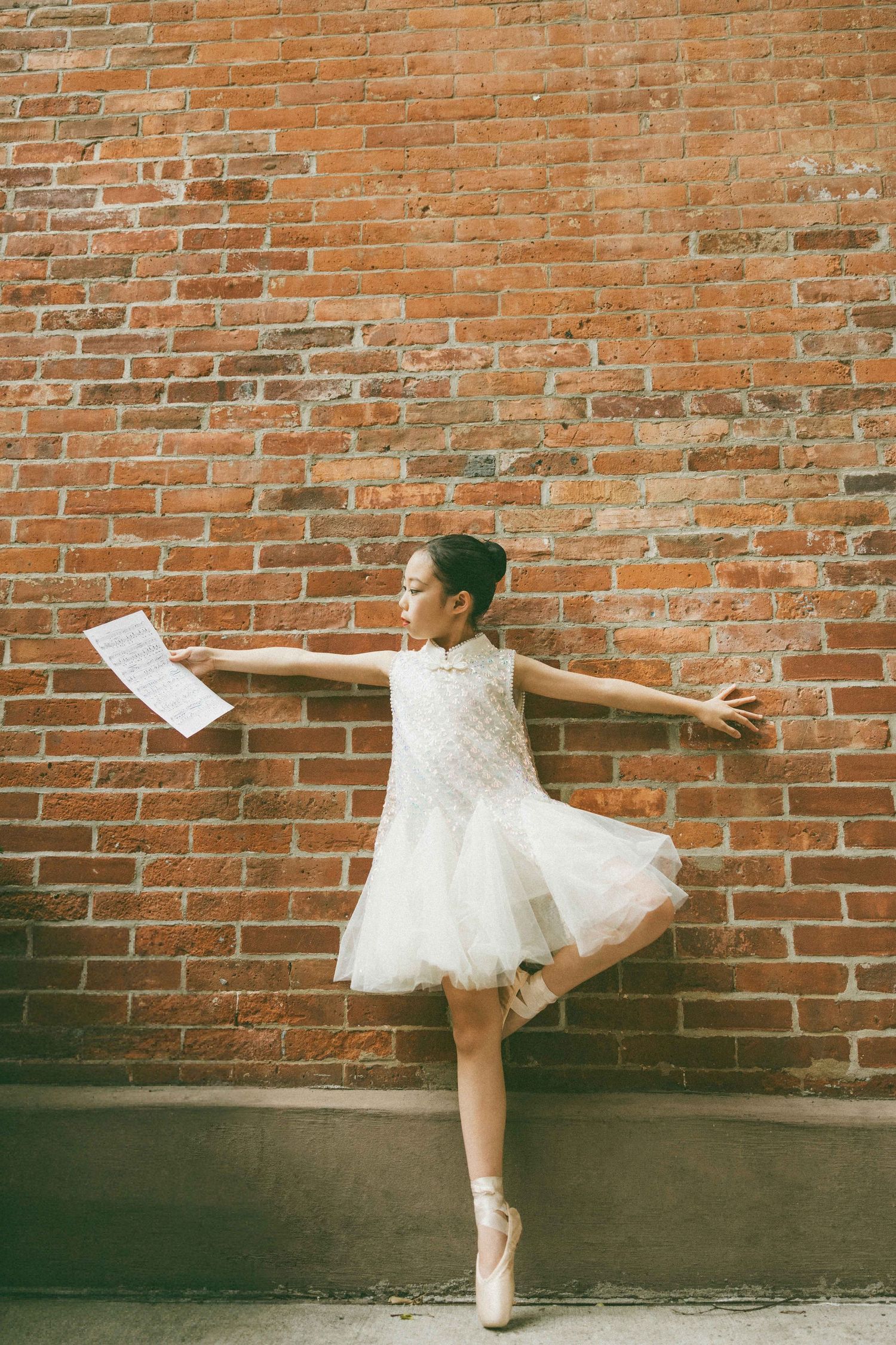 Young ballerina in white tutu dress poses on pointe shoes against a red brick wall outdoors.