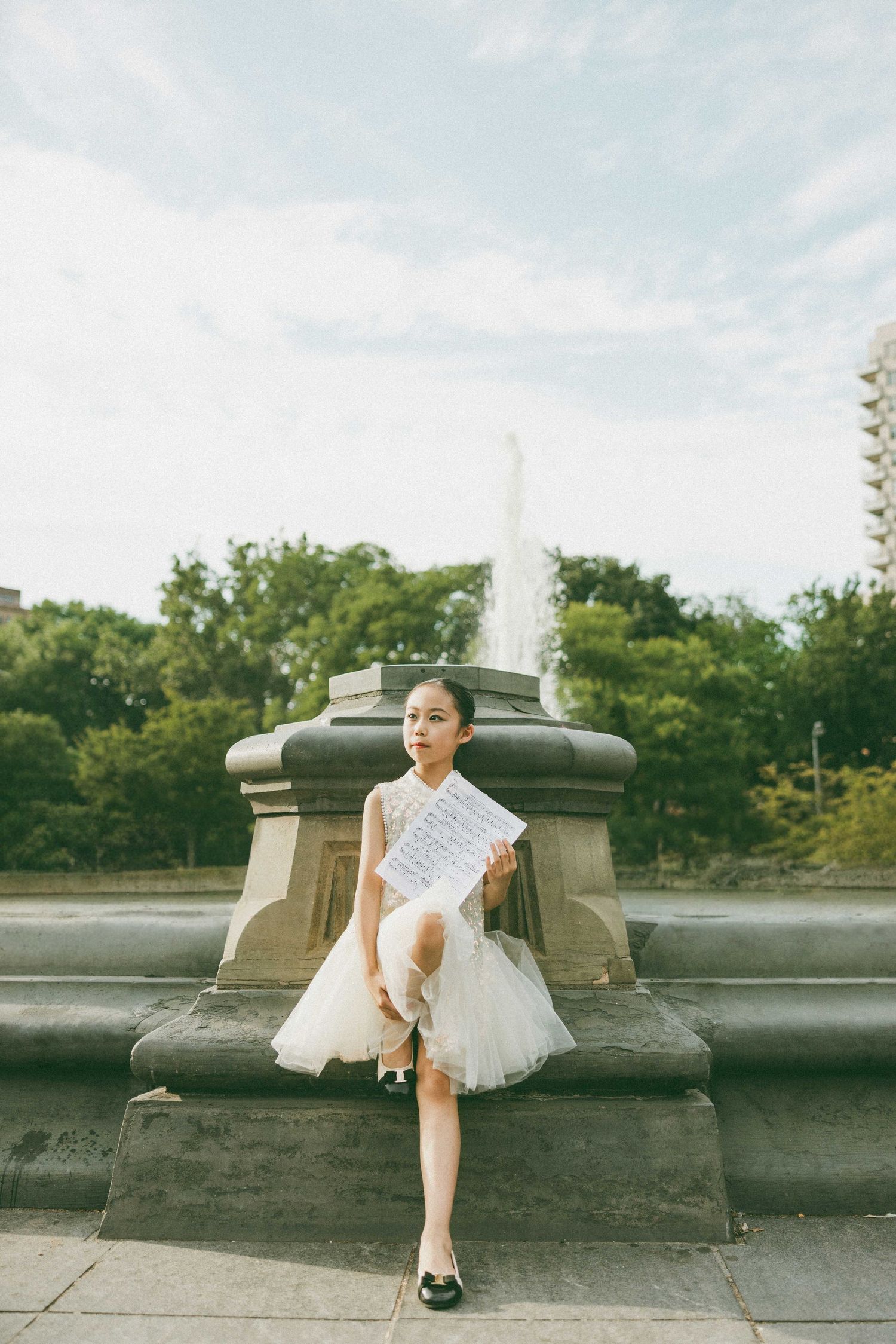 Young girl in white tulle dress sits at a stone fountain in a park, holding sheet music, with trees and sky behind her.