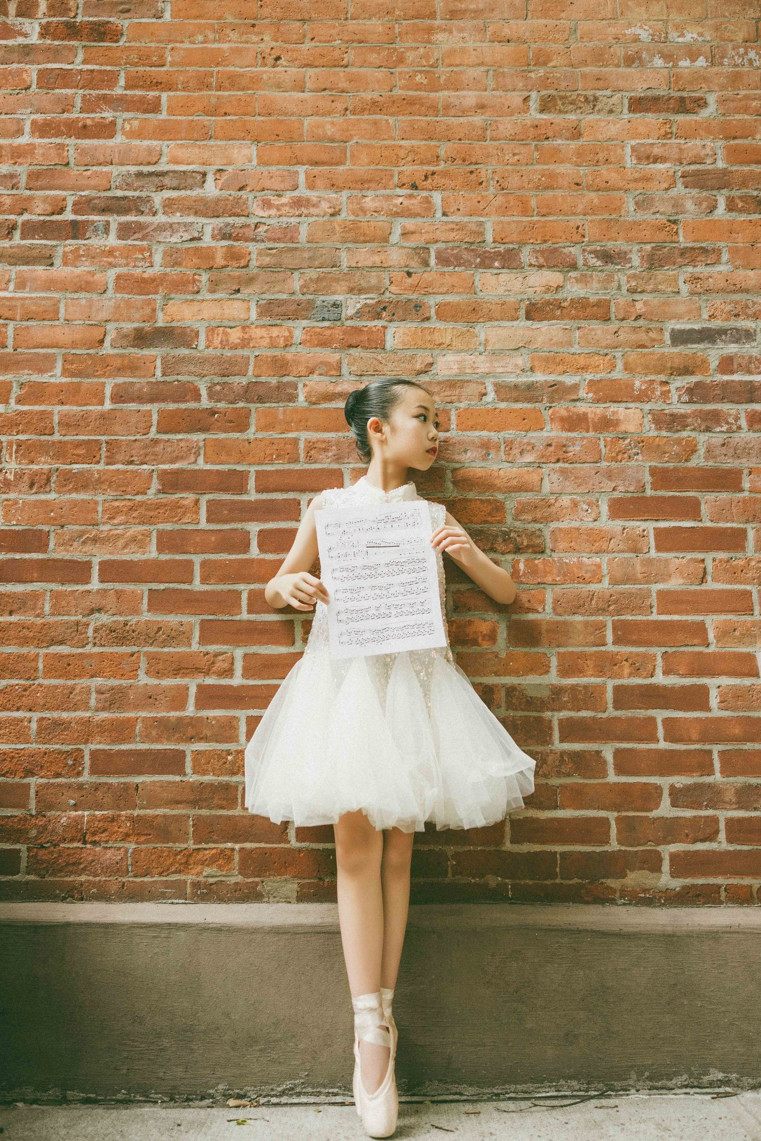 Young ballerina in white tutu holds sheet music against red brick wall, wearing pointe shoes.