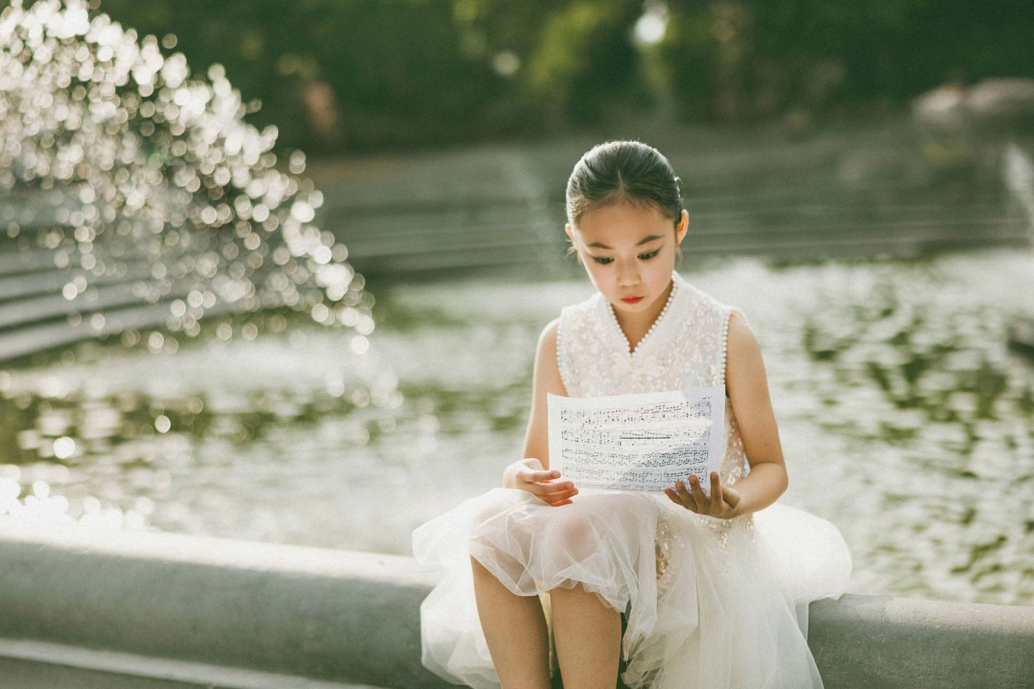 Young girl in white dress reads sheet music while sitting by a decorative fountain outdoors.
