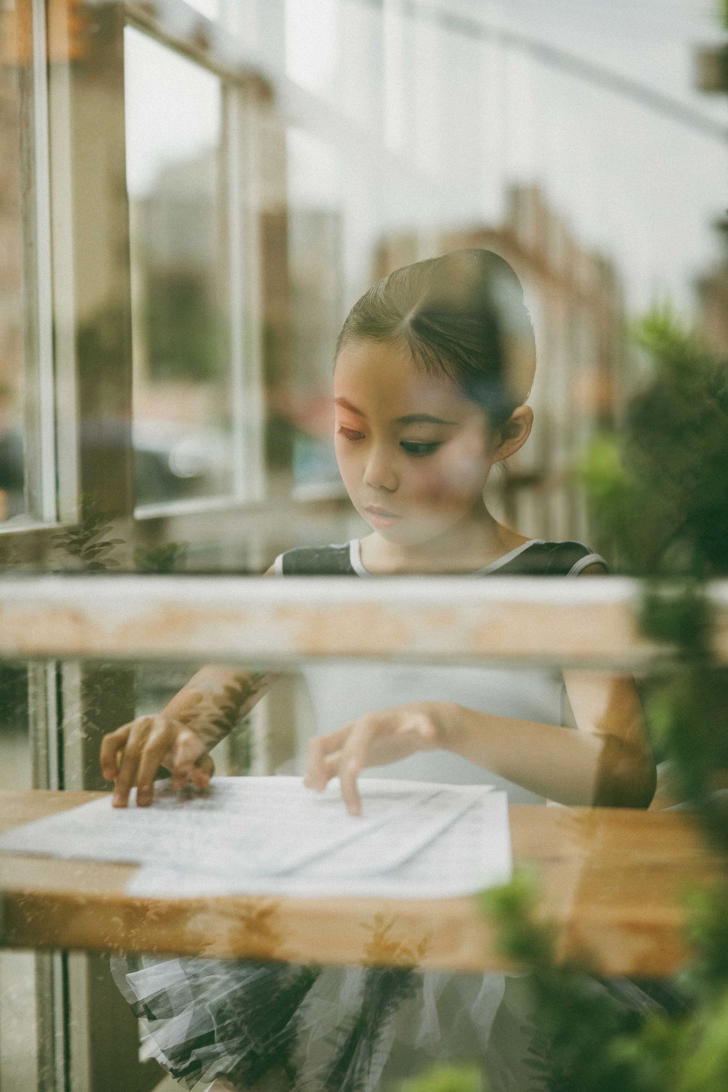 Young woman reading a document at a wooden table, viewed through a cafe window with city reflections.