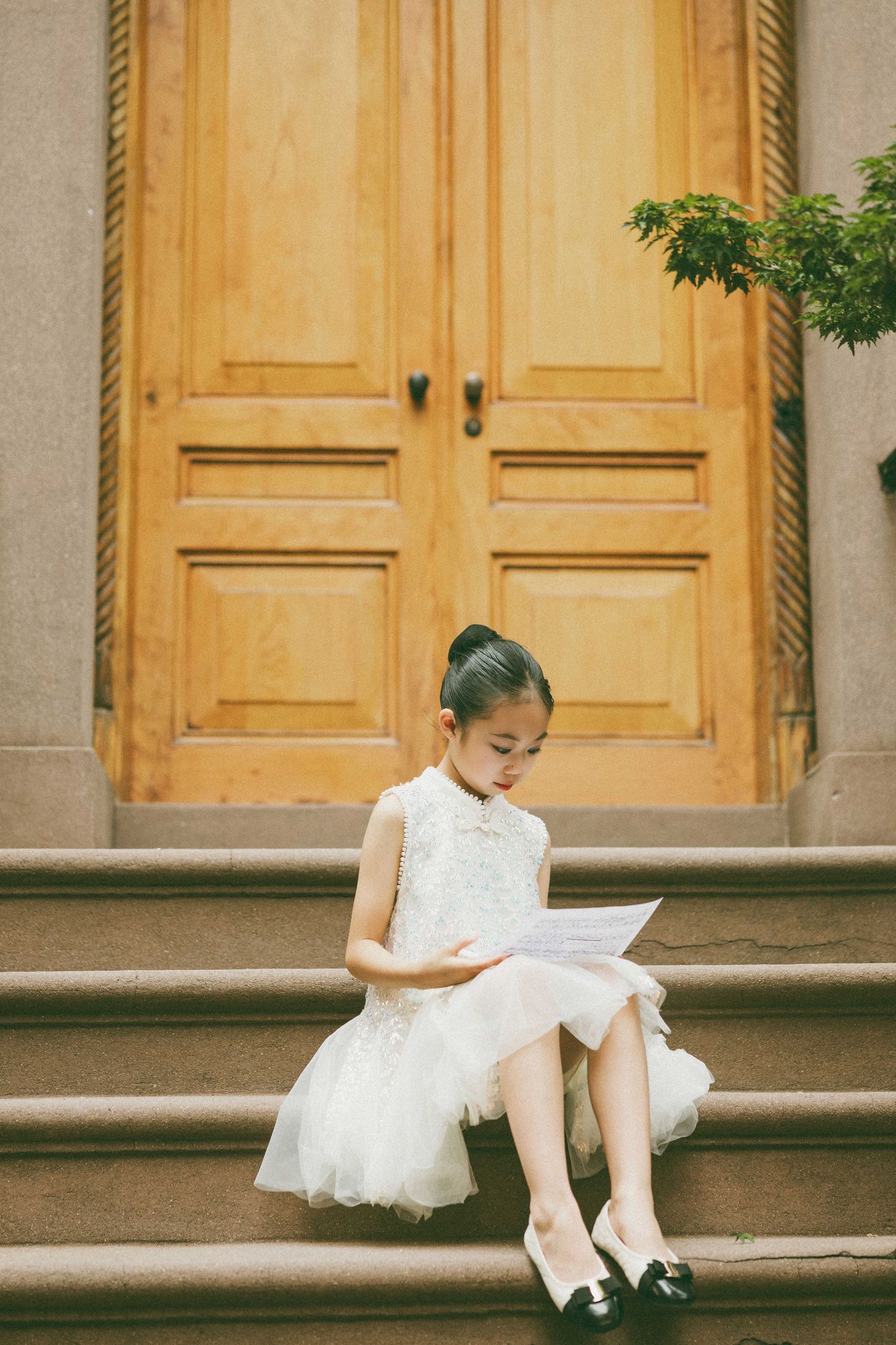 A young girl in a white lace dress sits on stone steps reading papers before large wooden doors.
