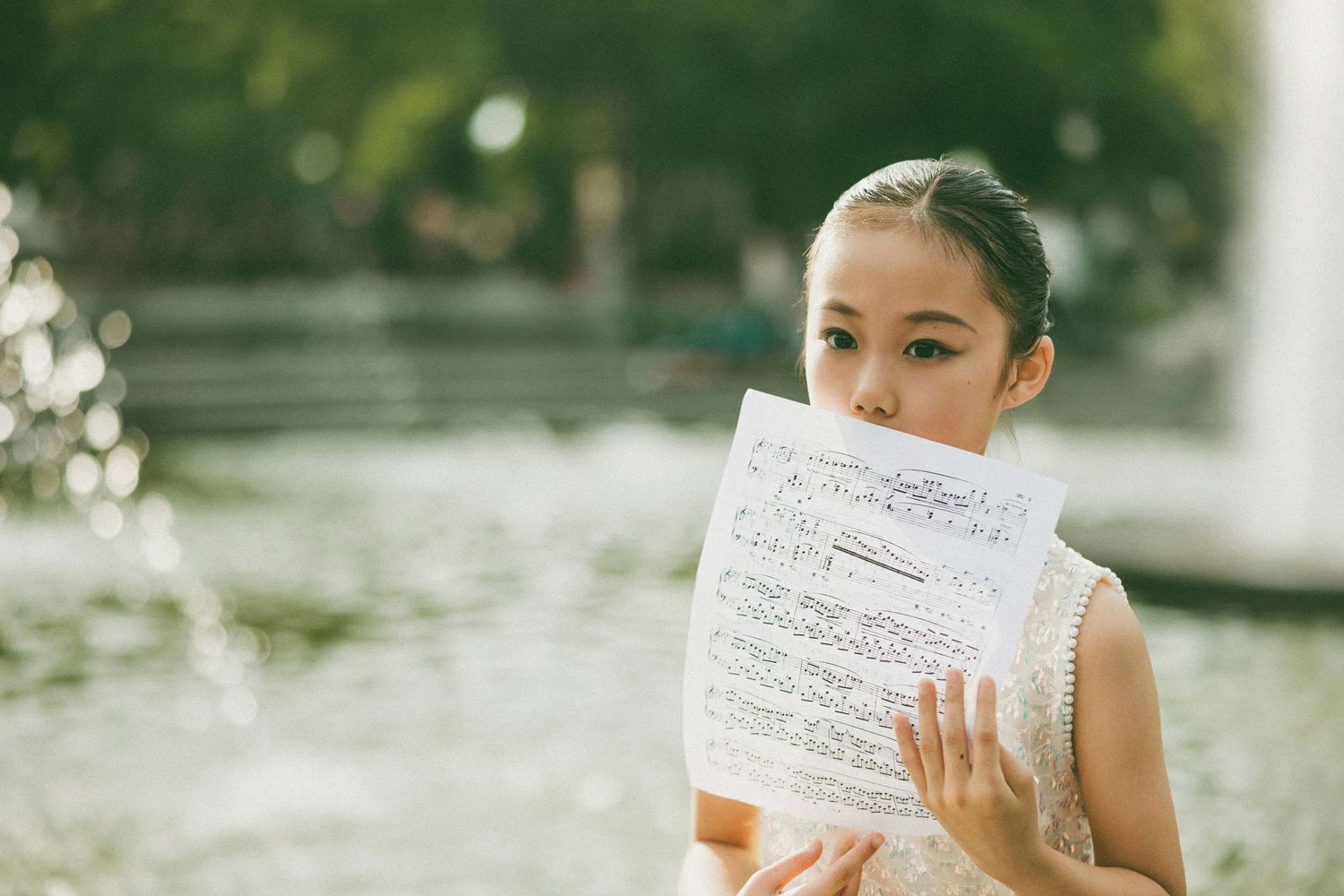 Young Asian girl holding sheet music in front of her face near a fountain outdoors.