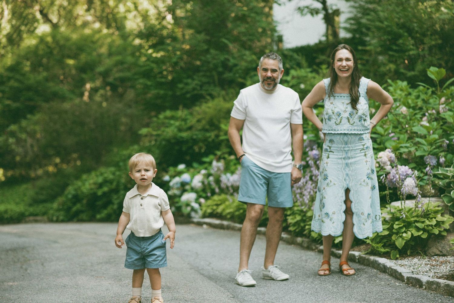 Family of three posing outdoors on a garden path surrounded by lush greenery and hydrangeas on a sunny day.