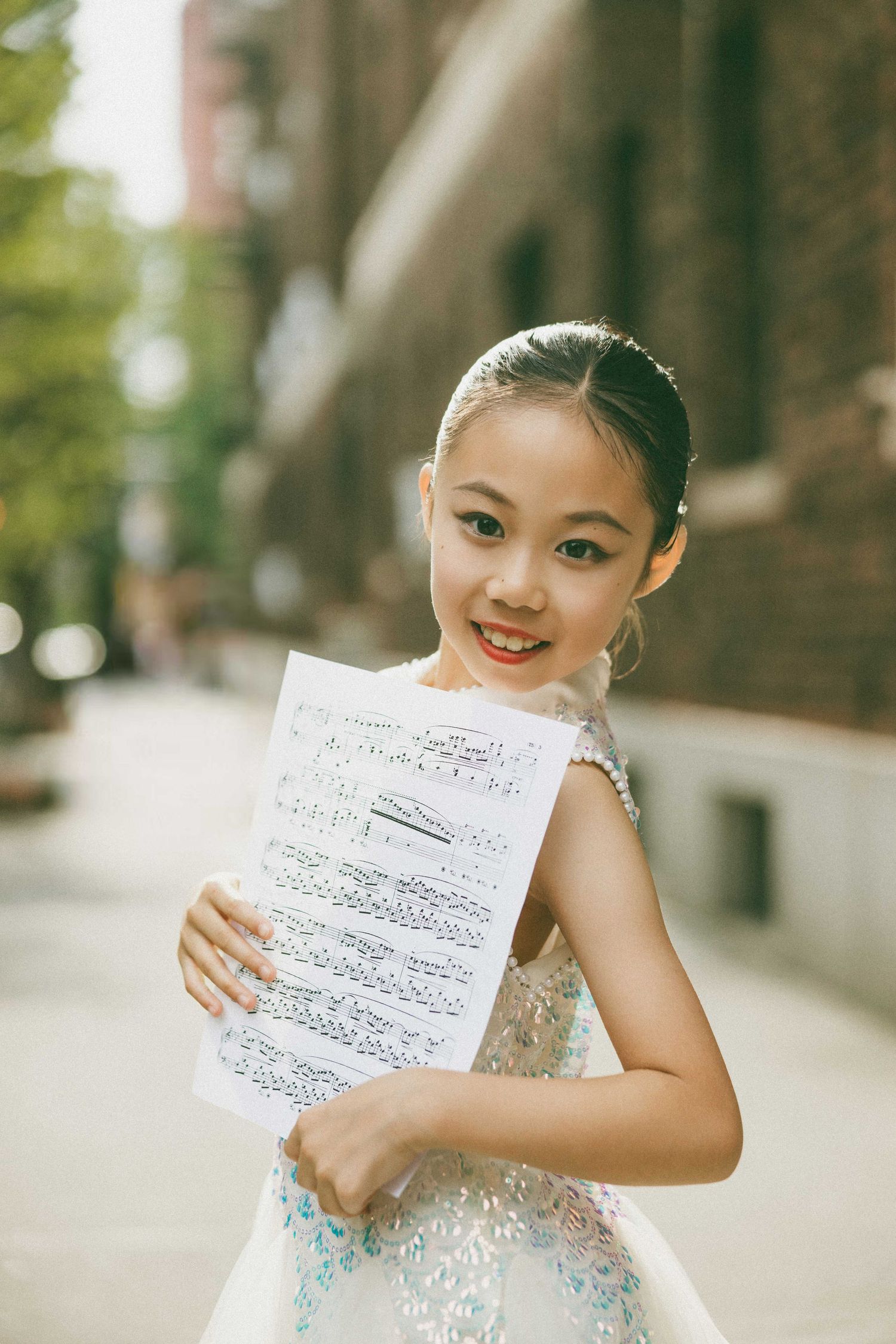 Young girl in sequined dress smiling while holding sheet music outdoors on a sunny urban street.