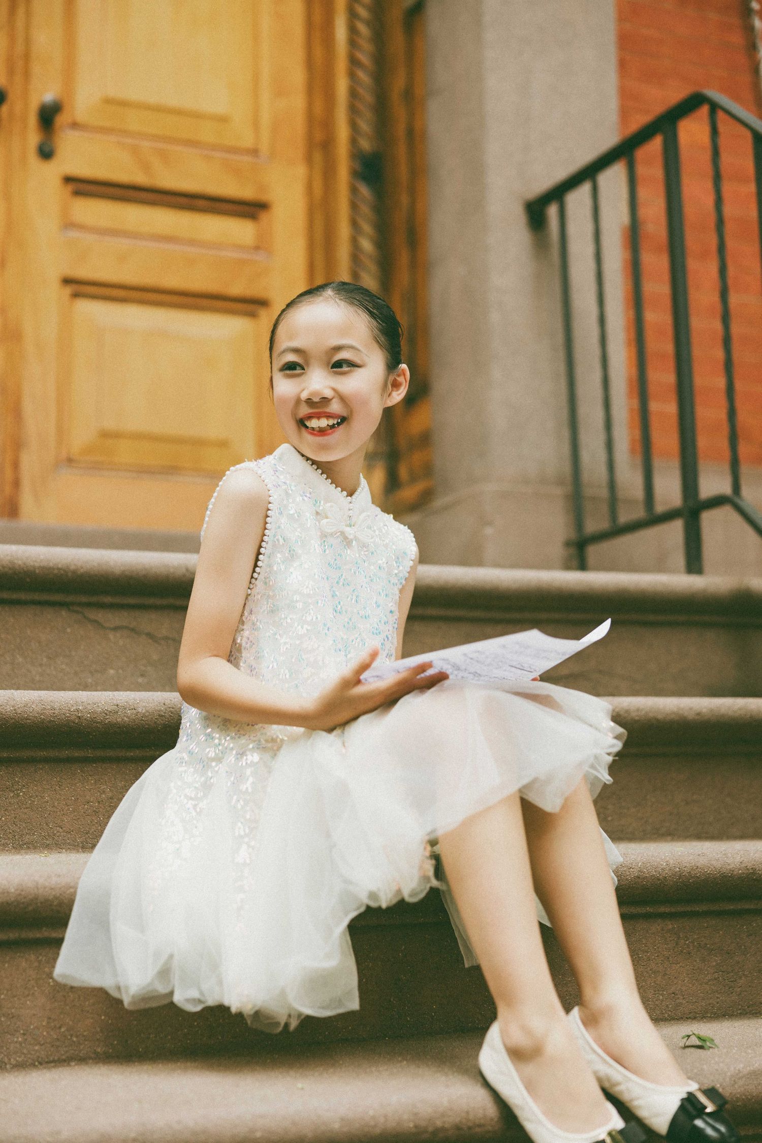 Smiling girl in white lace dress sits on brownstone steps holding sheet music, wearing flats.