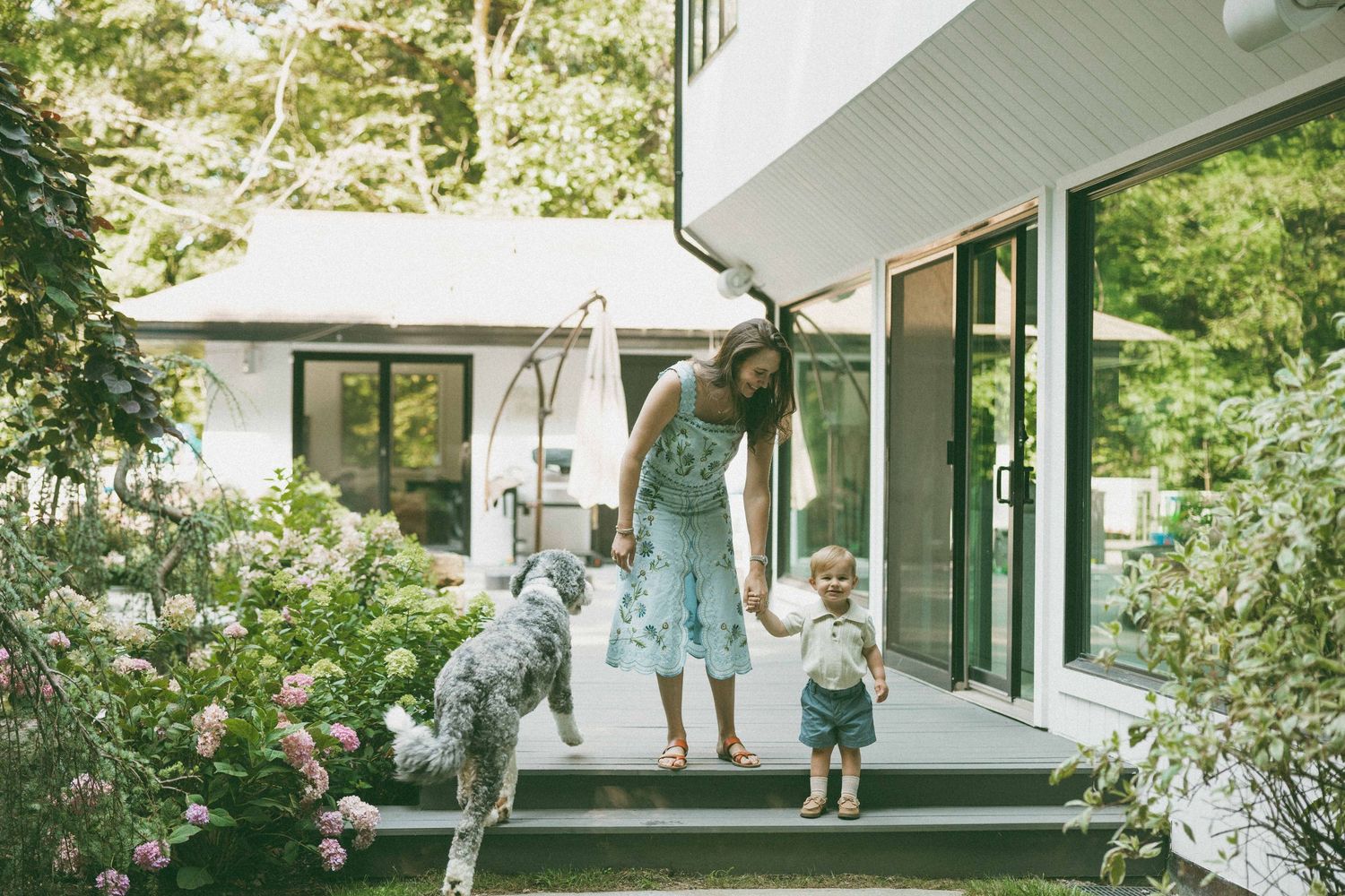 Woman in floral dress holds toddler's hand on patio steps beside dog, surrounded by lush garden and modern white home.