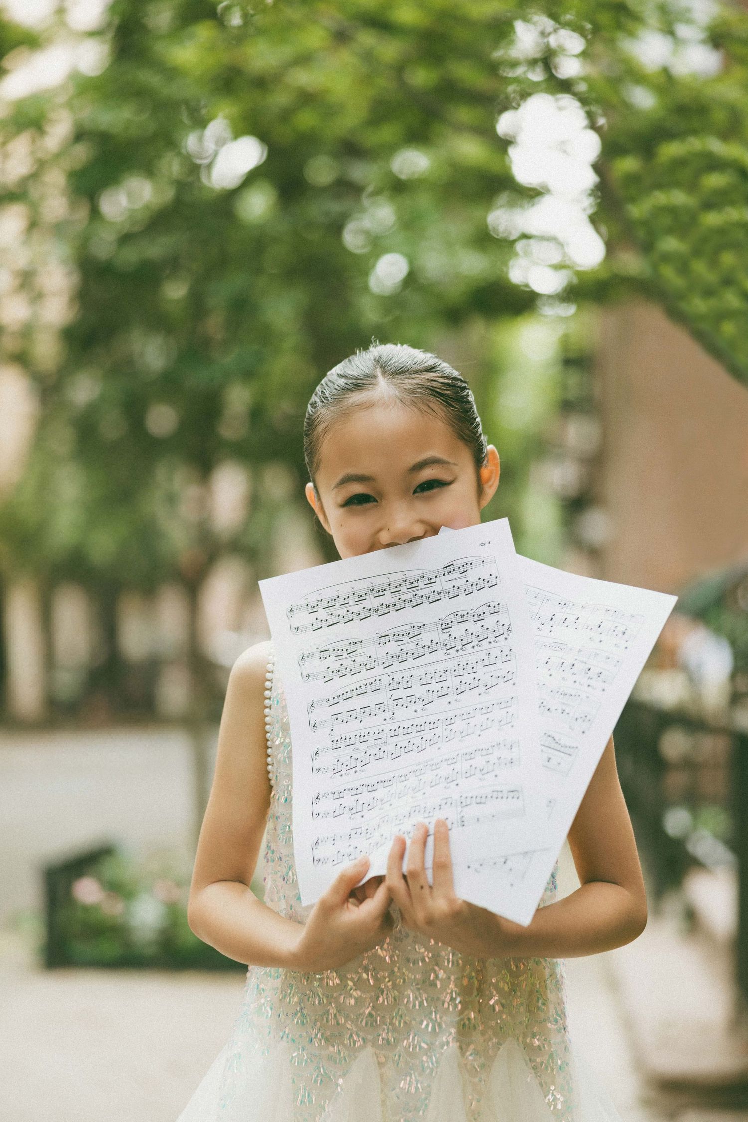 Young girl in white dress holding sheet music outdoors, smiling joyfully among green trees.