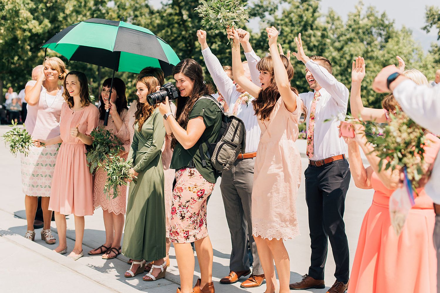Wedding guests celebrate with raised hands under a green umbrella outdoors.