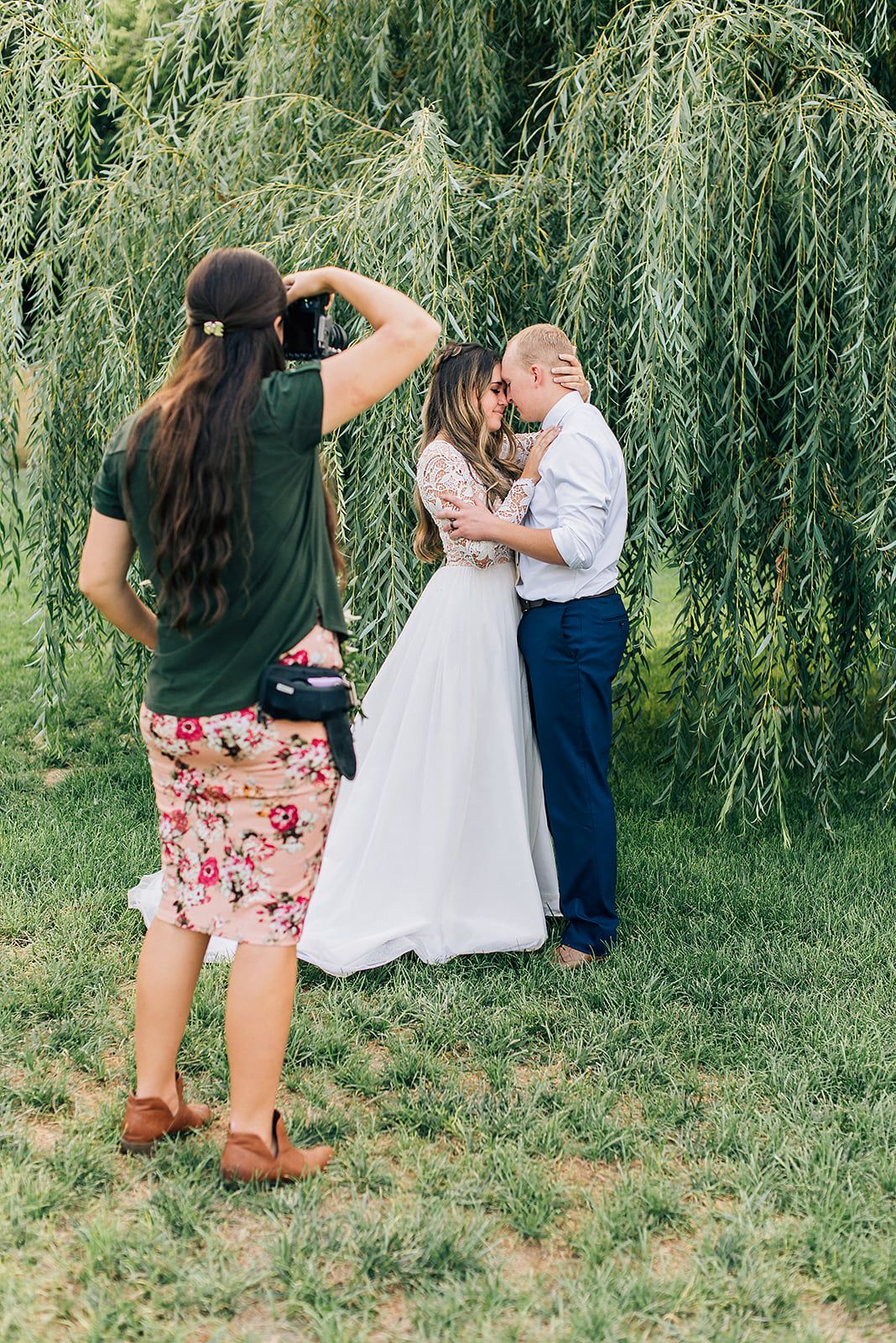 Photographer captures romantic moment under willow tree during wedding photoshoot.