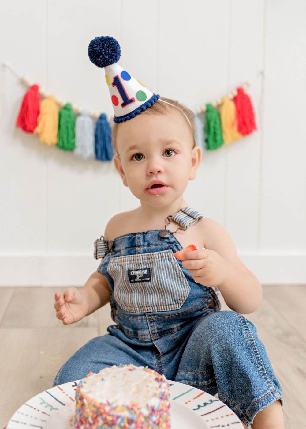 A baby in denim overalls sits on a white floor with colorful party decorations and a smash cake during a birthday celebration.