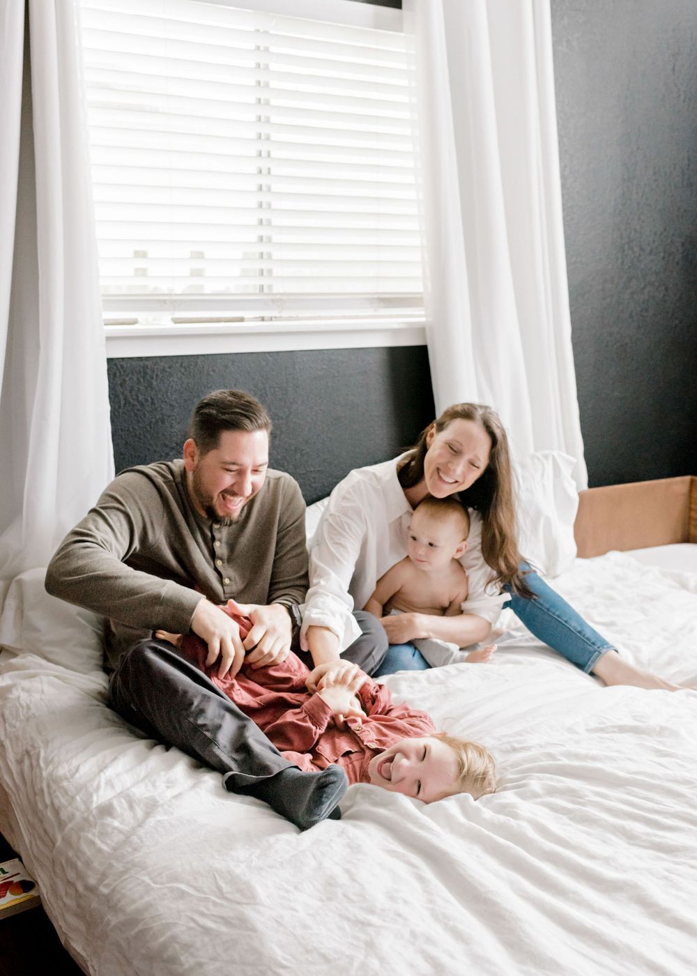 A family enjoys playful moments together while lounging on a white bed with natural light streaming through windows.