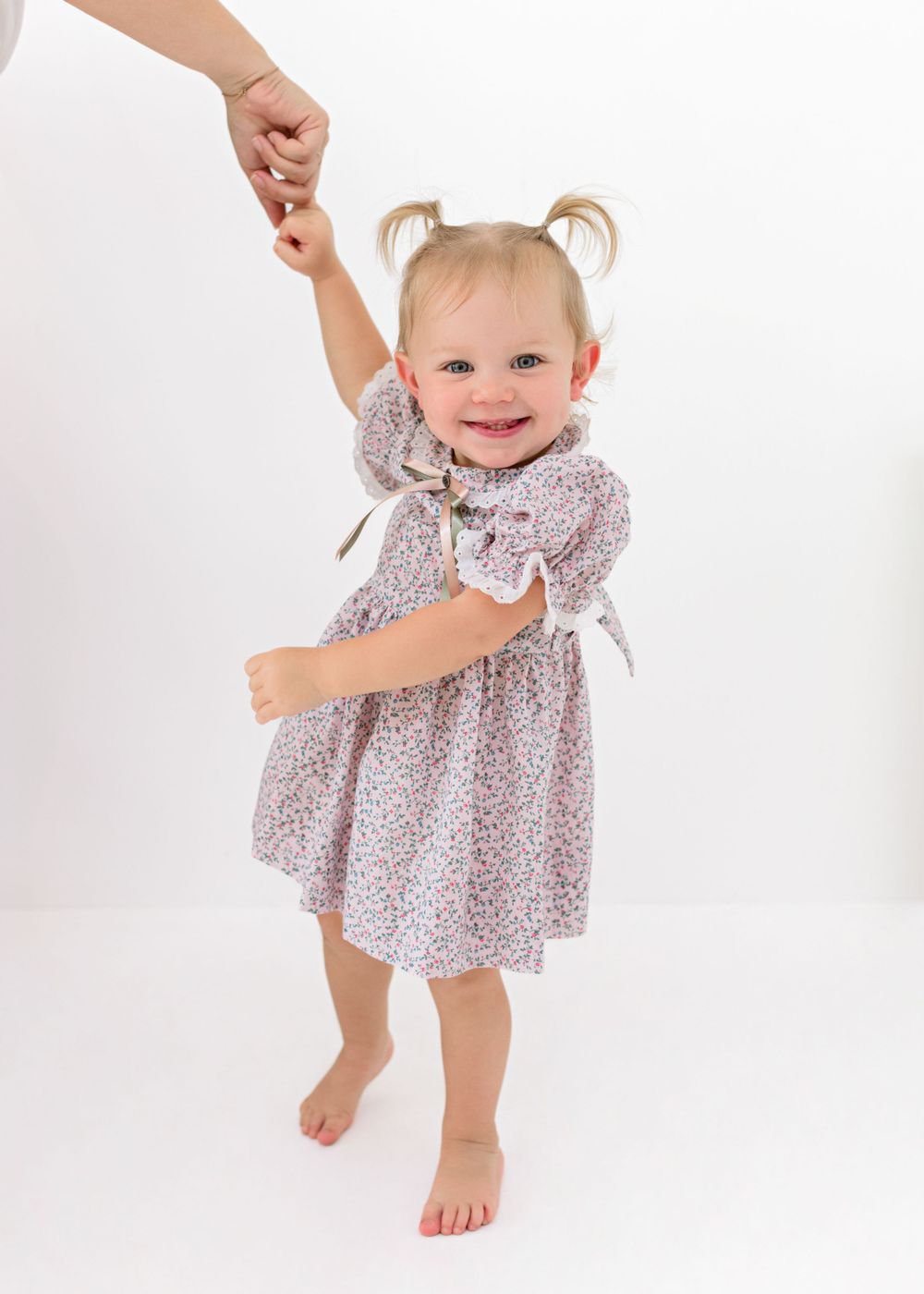 Smiling toddler in floral dress dancing while holding adult's hand against white background.