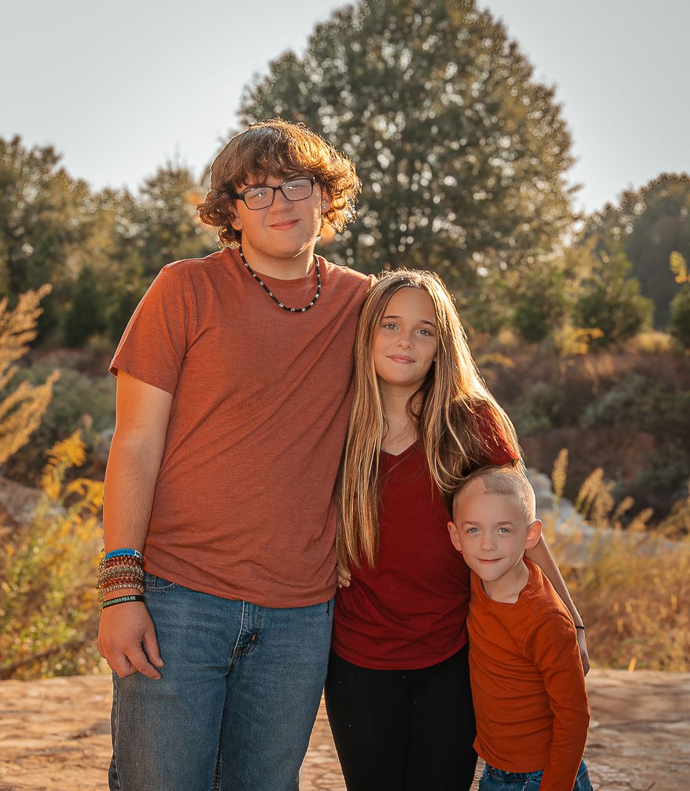 Three young siblings in red and orange shirts pose together outdoors during golden hour in a natural setting.