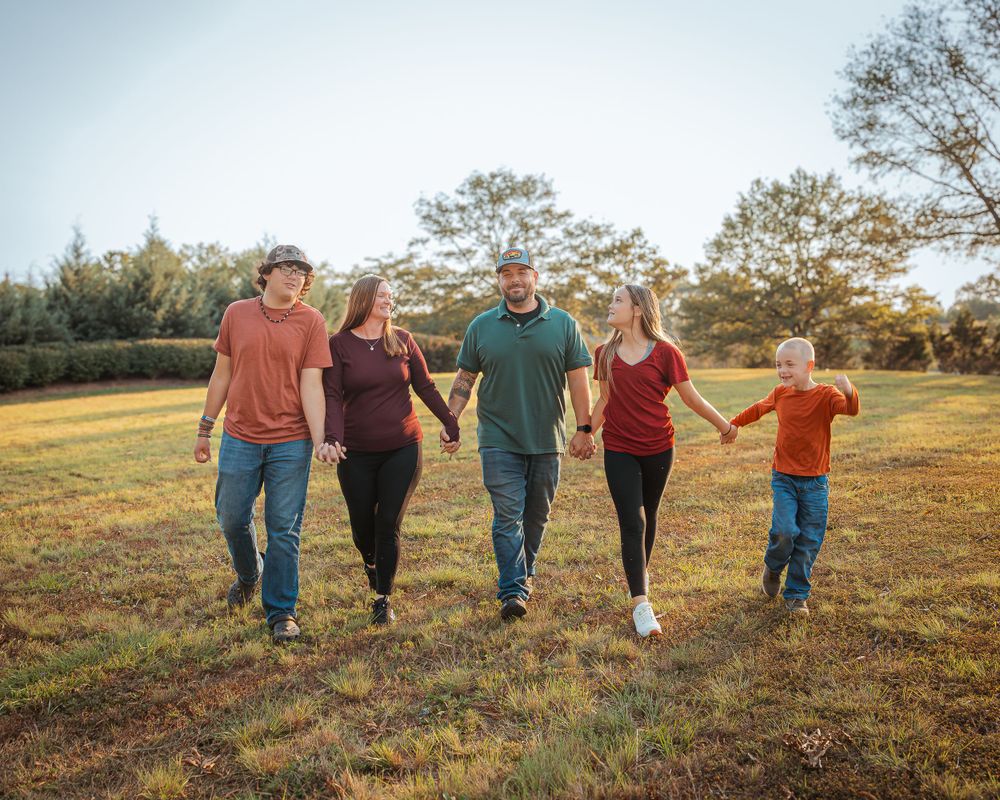 Five people walking together and holding hands across a sunny grassy field with trees in the background.