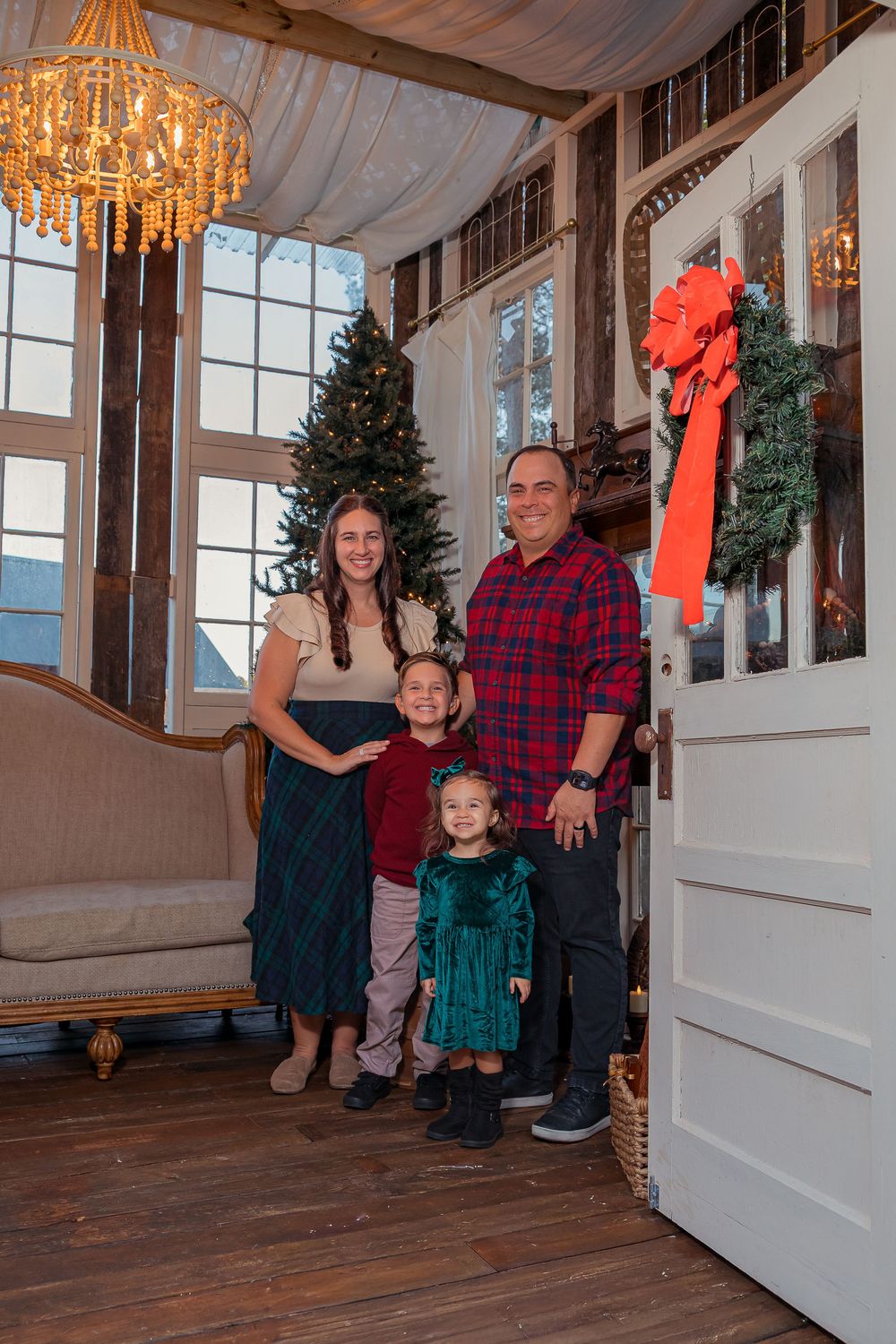 A family poses together in a festive holiday setting with Christmas trees and chandeliers in an elegant room.