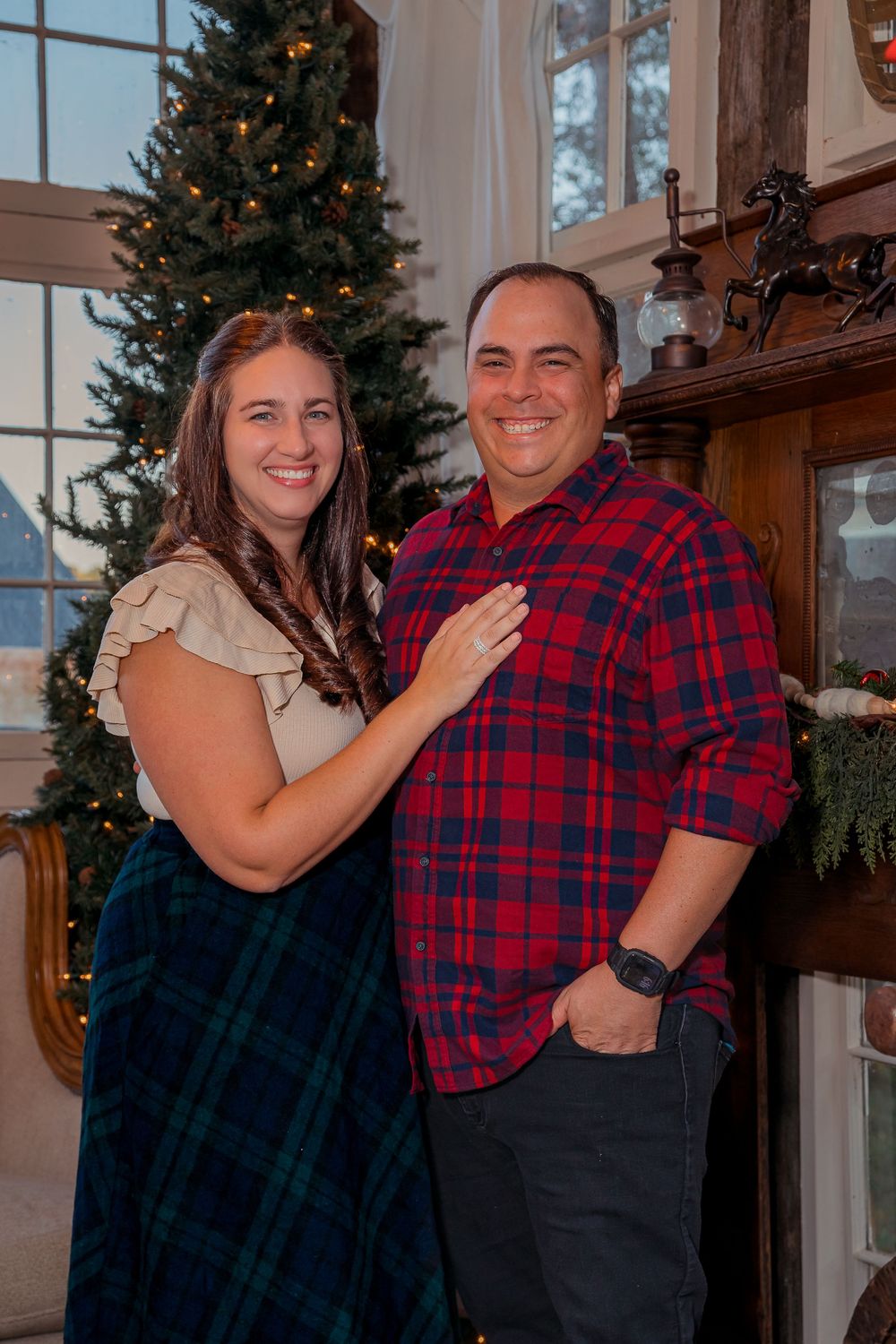 A couple poses together in front of a decorated Christmas tree in a cozy indoor setting wearing red and blue attire.
