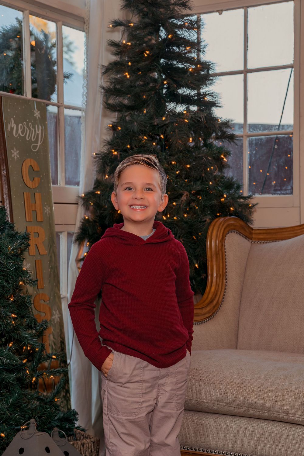 A child in a burgundy sweater poses and smiles in front of a decorated Christmas tree and beige couch.