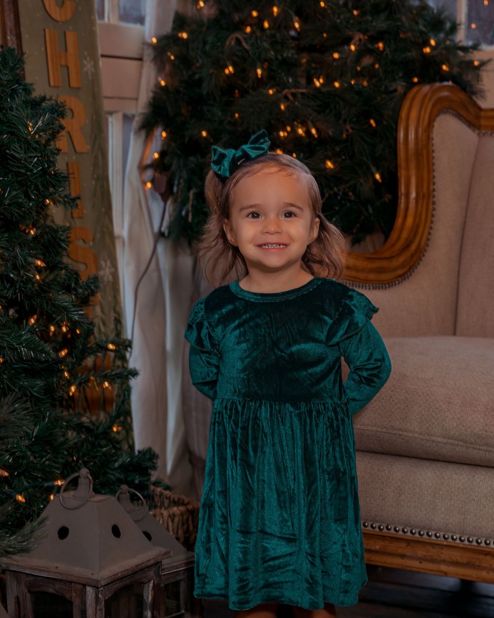 Child in emerald green velvet dress poses near decorated Christmas tree with warm holiday lighting.
