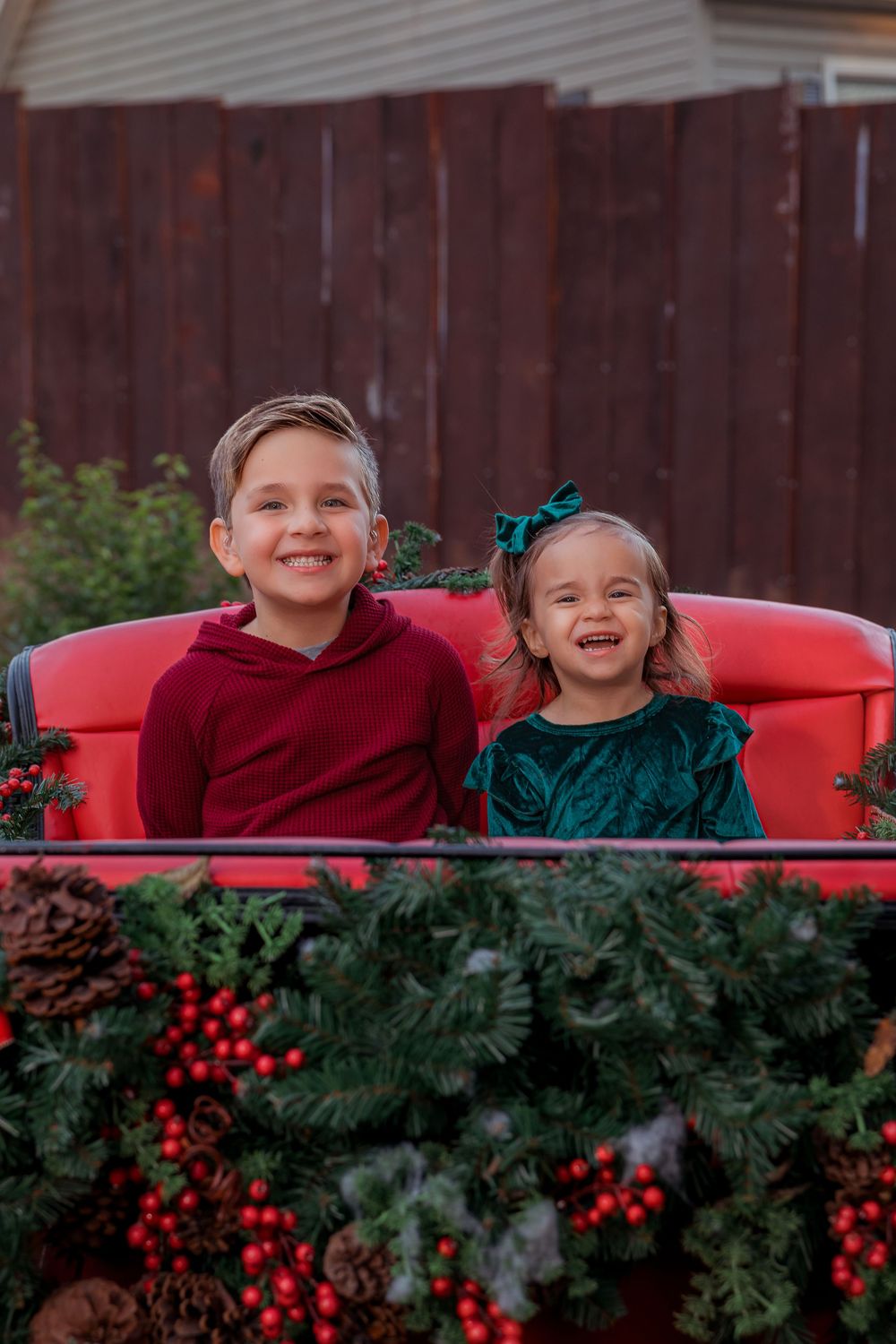 Children peek over a red truck bed decorated with festive holiday greenery and bright red berries.